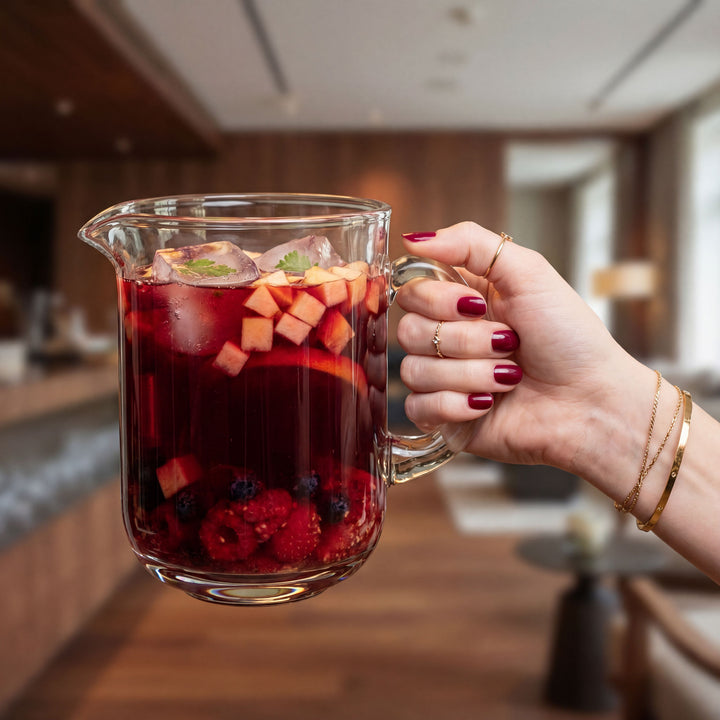 Hand holding a glass pitcher filled with a red beverage and fruit in a kitchen setting