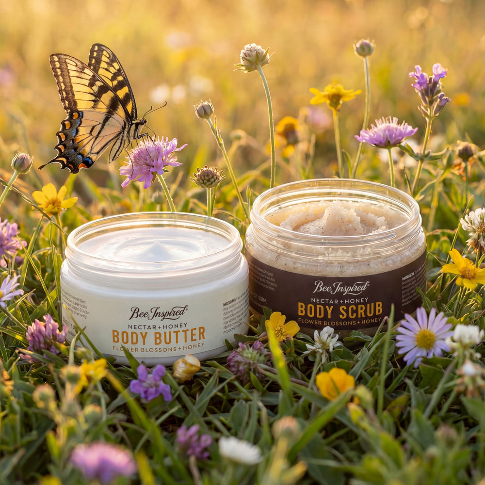 Two jars of 'Bee Inspired' body butter and scrub on a grassy field with flowers and a butterfly.