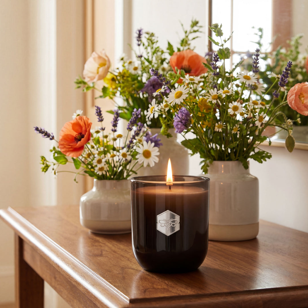 Candle with a hexagonal logo on a wooden surface with floral arrangements in the background.