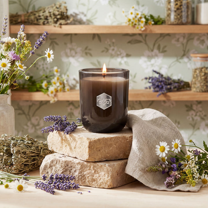 Black candle with a logo on a stone surface surrounded by flowers and herbs.