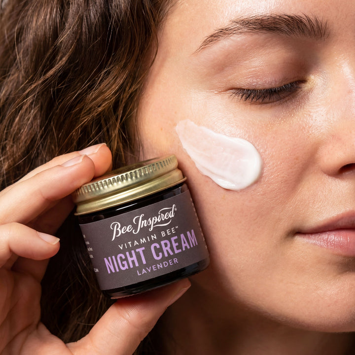 Person applying night cream to their face with a jar labeled 'Bee Inspired Vitamin Bee Night Cream Lavender'.