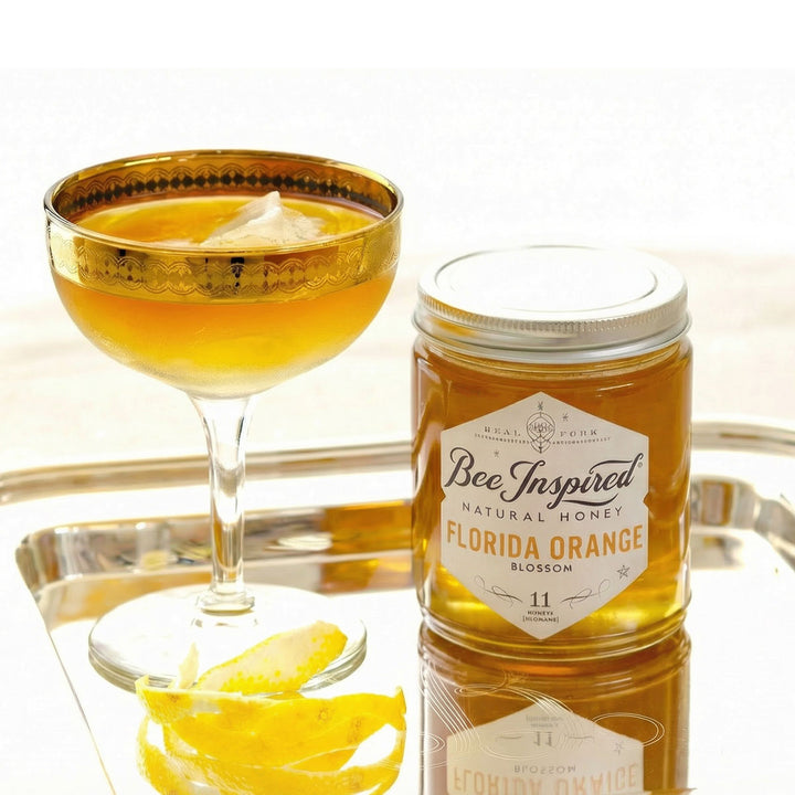 Glass of bourbon and honey cocktail with a jar of Bee Inspired Florida Orange honey on a reflective surface.