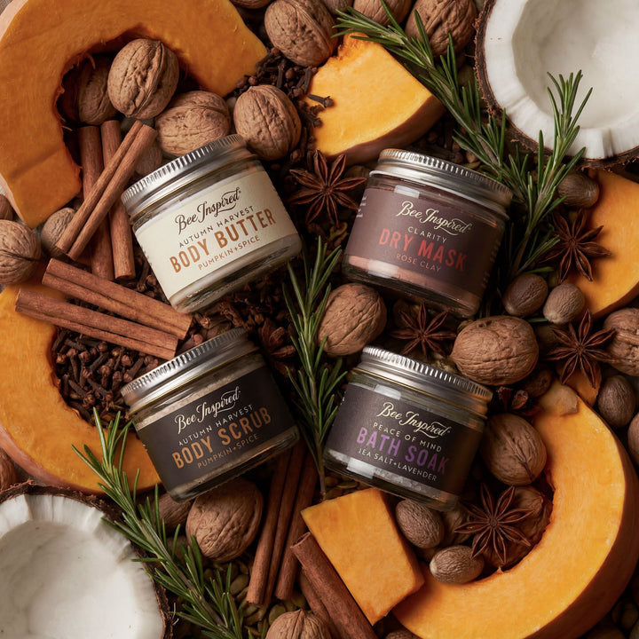Four jars of Bee Inspired body care products surrounded by autumnal elements like pumpkins, nuts, and spices.