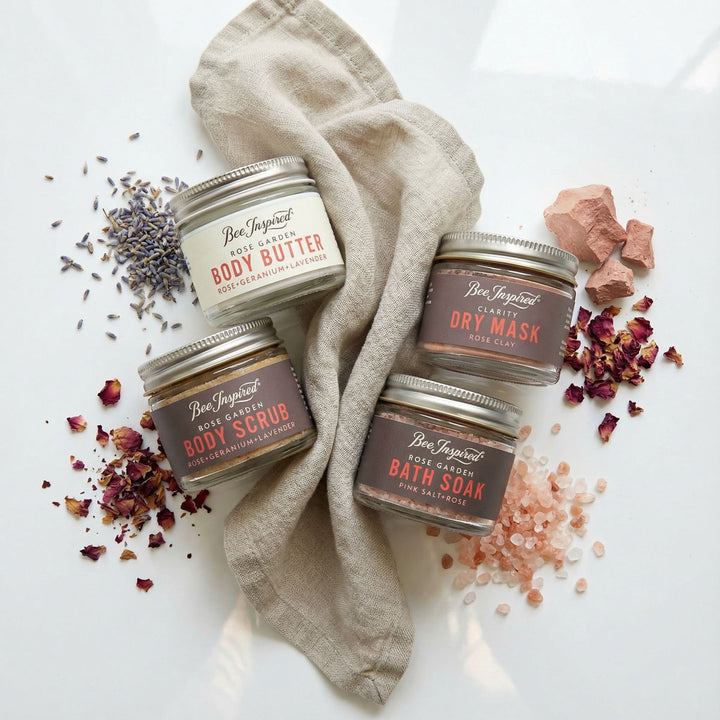 Four jars of Bee Inspired body care products on a white background with lavender, rose petals, and a towel.