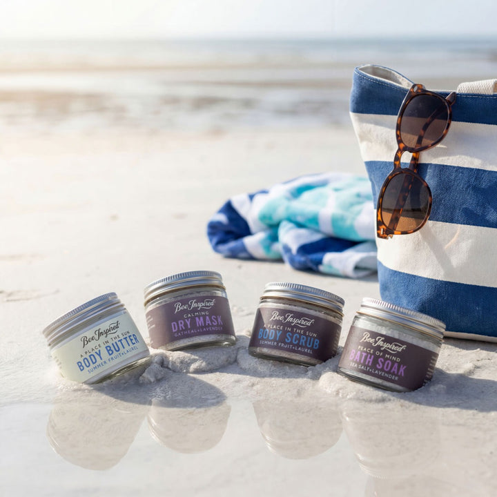 Four jars of Bee Inspired products on a sandy beach with a striped bag and sunglasses.