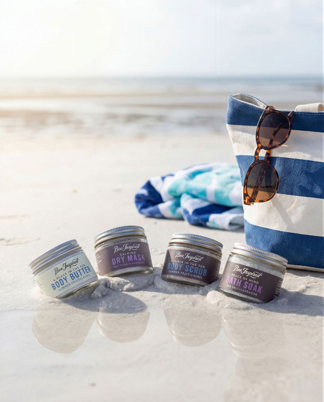 Four jars of body care products on a sandy beach with a towel and sunglasses in the background.