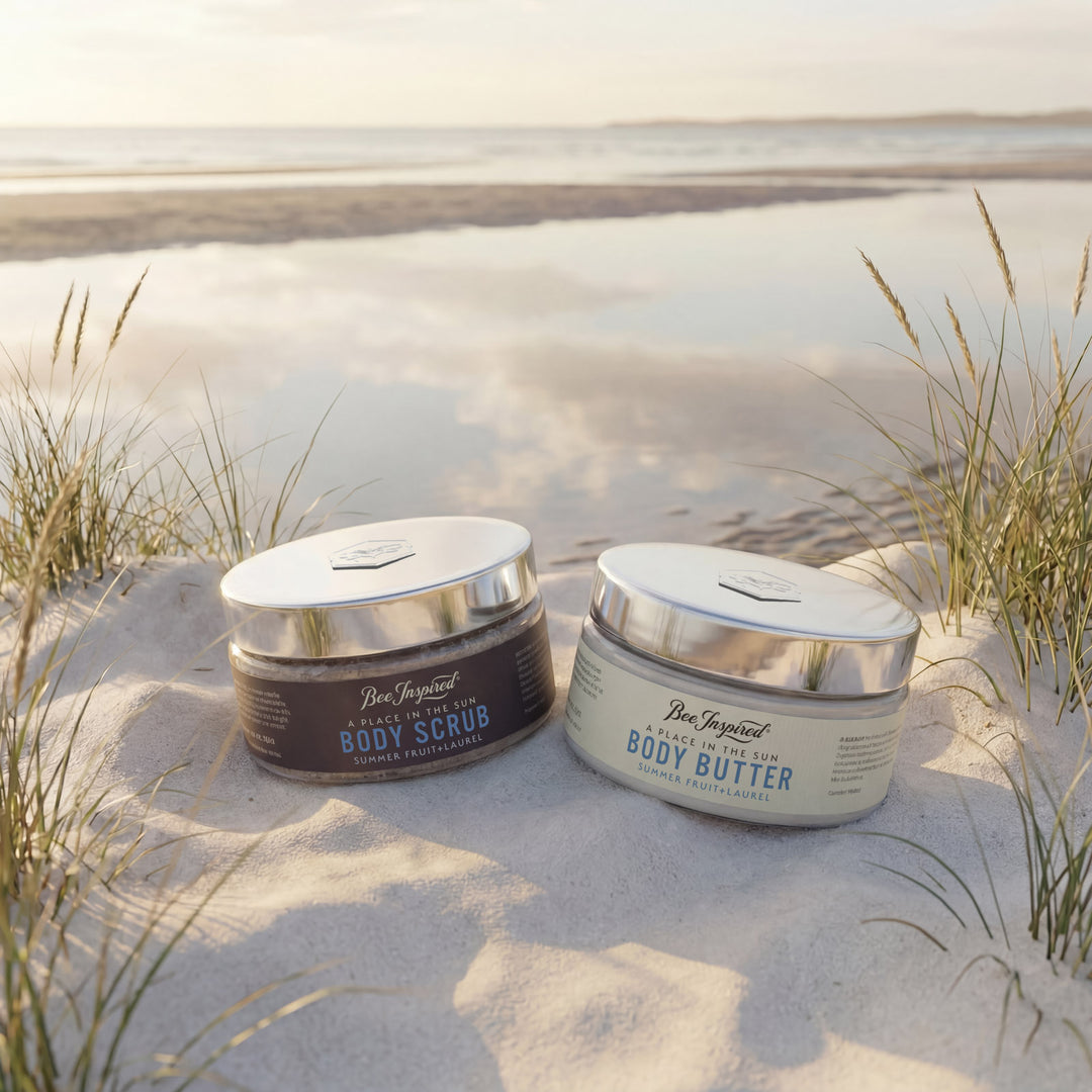 Two jars of Bee Inspired Body Scrub and Body Butter on a sandy beach with grasses.