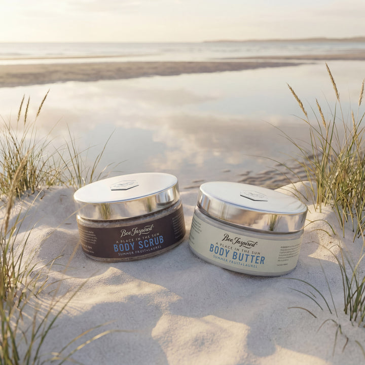 Two jars of Bee Inspired Body Scrub and Body Butter on a sandy beach with grasses.