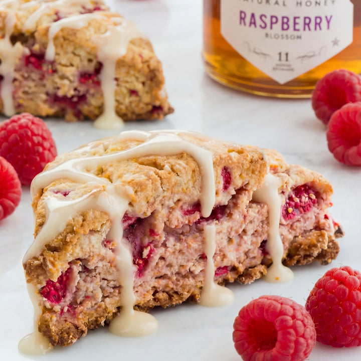 Raspberry scone with a drizzle of cream cheese glaze, surrounded by fresh raspberries and a bottle of raspberry-flavored honey.