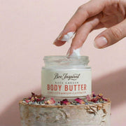 Hand applying body butter from a jar labeled 'Bee Inspired Rose Garden' on a pink background