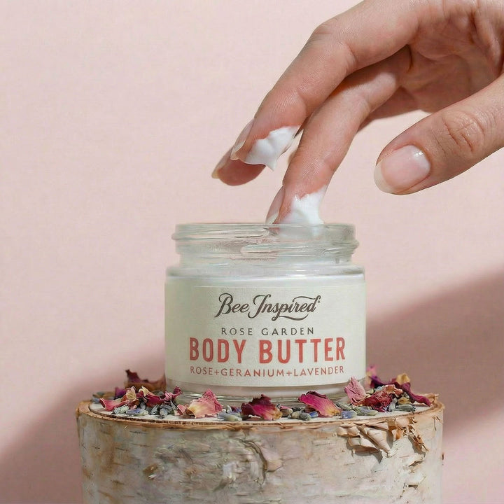 Hand applying body butter from a jar labeled 'Bee Inspired Rose Garden' on a pink background