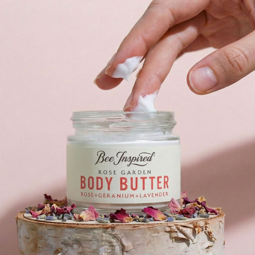 Hand applying body butter from a jar labeled 'Bee Inspired Rose Garden Body Butter' on a pink background.