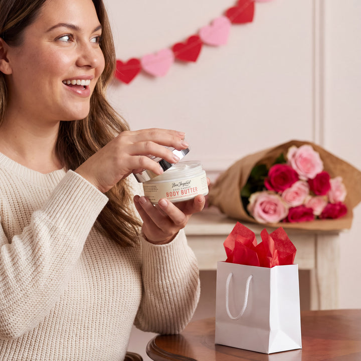 Woman holding a jar of body butter with a bouquet of roses and gift bag in the background