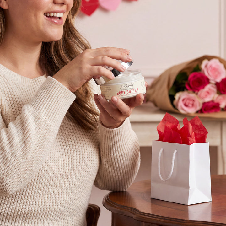 Woman holding a jar of body butter with a bouquet of flowers and gift bag in the background