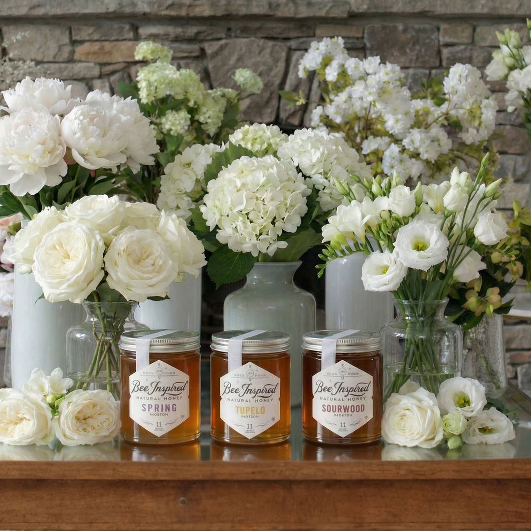 Jars of Bee Inspired Royal honey with floral bouquets on a wooden surface against a stone wall.