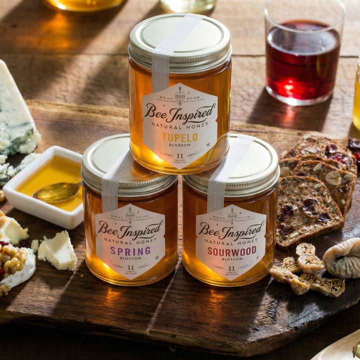 Three jars of 'Bee Inspired' royale honey on a wooden board with cheese, fruit, crackers and drinks.