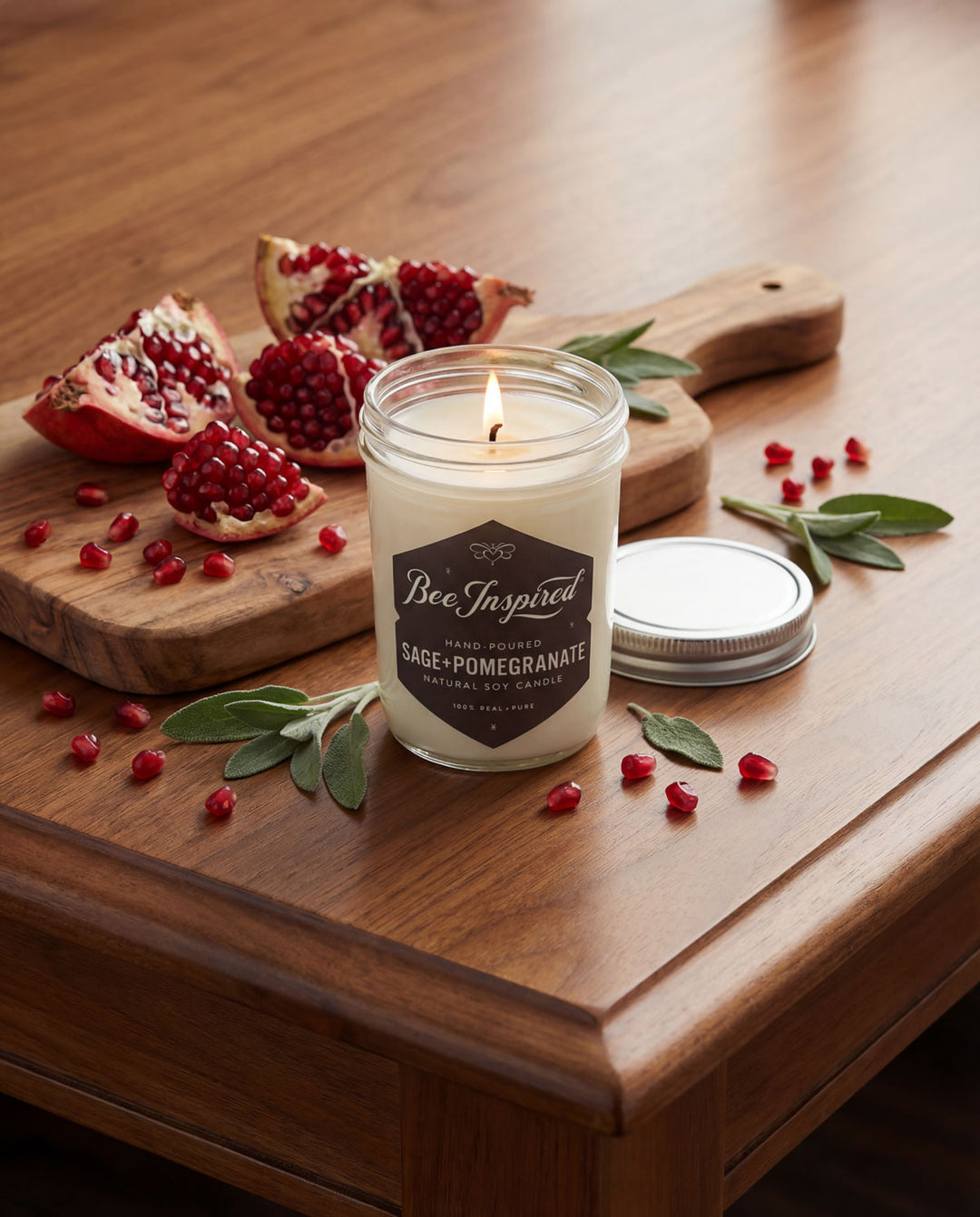 Candle with 'Bee Inspired' branding on a wooden table with pomegranate seeds and leaves.