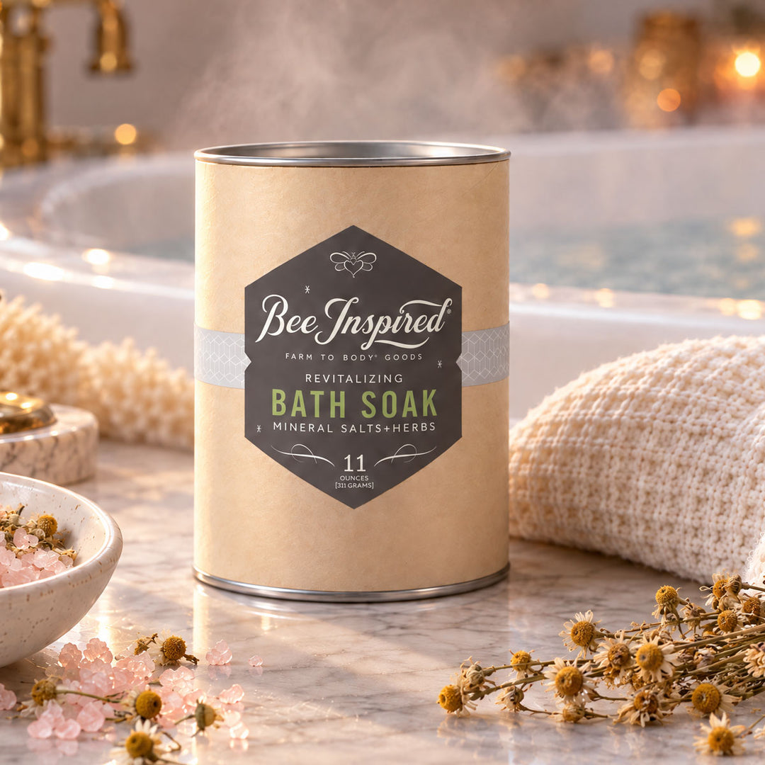 A canister of Bee Inspired Revitalizing Bath Soak sits on a marble countertop in a bathroom setting, surrounded by a beige knit towel, a bowl of pink bath salts, and small dried flowers.