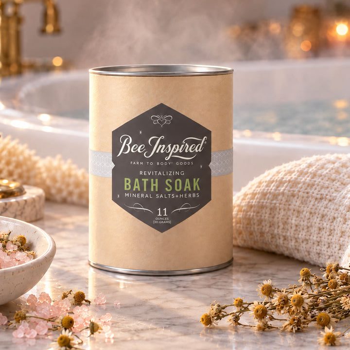 A canister of Bee Inspired Revitalizing Bath Soak sits on a marble countertop in a bathroom setting, surrounded by a beige knit towel, a bowl of pink bath salts, and small dried flowers.