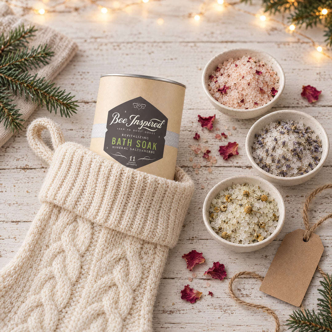 A kraft paper bath soak tin labeled Bee Inspired Bath Soak sits in a cream cable-knit stocking on a whitewashed wooden surface. Three small bowls of bath salts with dried botanicals are arranged beside it, along with scattered rose petals, evergreen branches, and warm fairy lights.