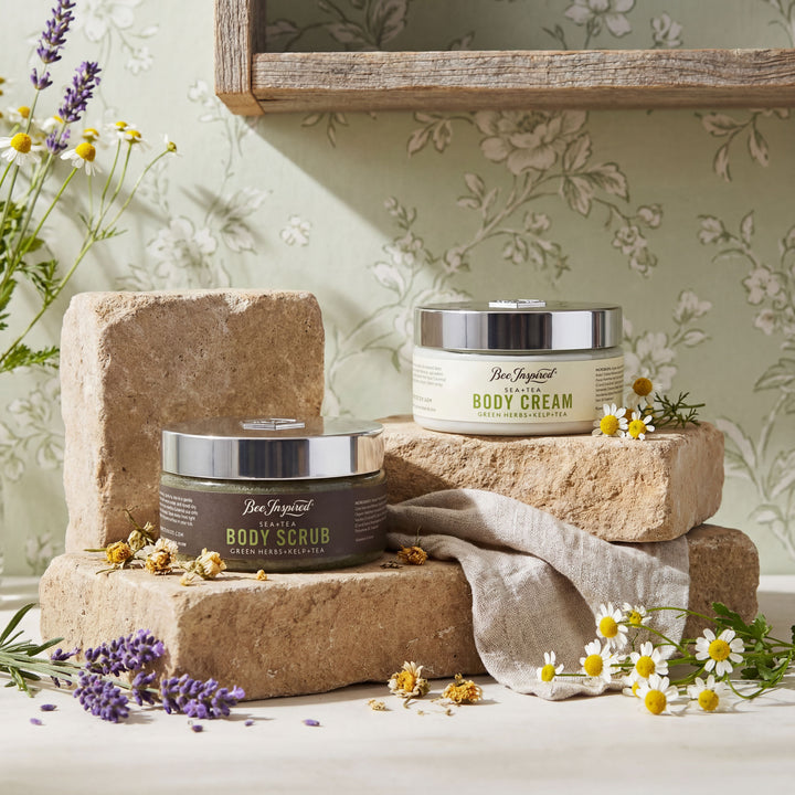 Two jars of body cream and scrub on stone blocks with flowers and a towel in the background.