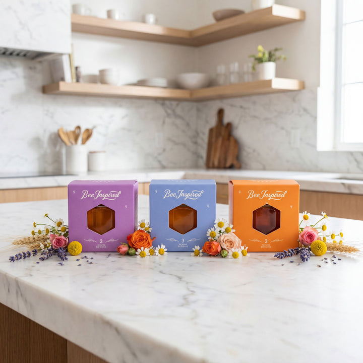 Three 'Bee Inspired' product boxes on a kitchen counter with flowers and shelves in the background.