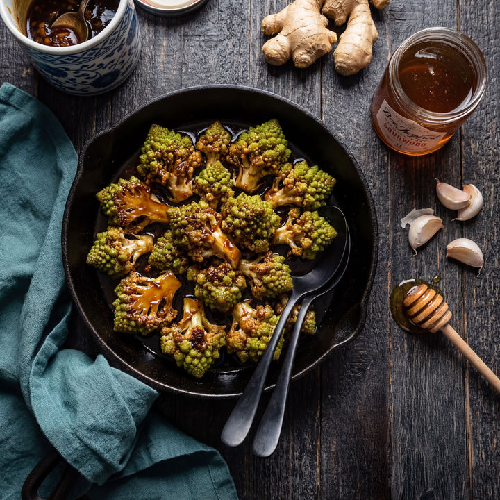 Roasted cauliflower in a black bowl with a jar of honey and ginger on a wooden table.
