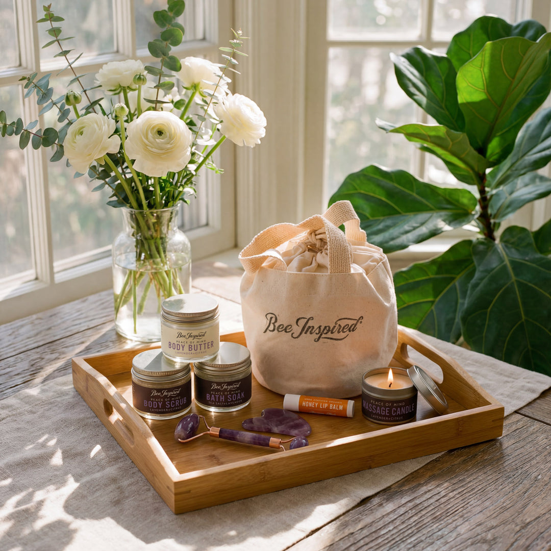 Bathroom set with body butter, candles, and a tote bag on a wooden tray with flowers and plants in the background.
