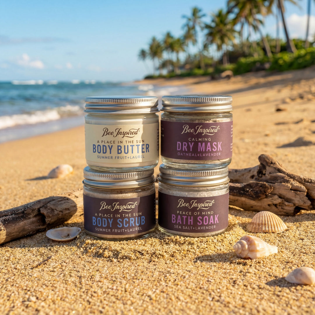 Four containers of Bee Inspired products on a sandy beach with ocean and palm trees in the background.