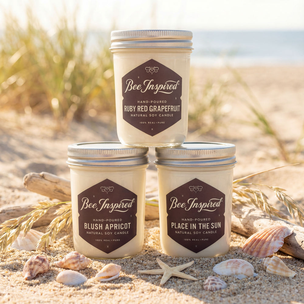 Three 'Bee Inspired' candles stacked on a sandy beach with seashells and driftwood.