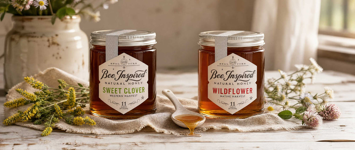 Two glass jars of honey on a rustic white wooden table, labeled Bee Inspired Natural Honey Sweet Clover and Wildflower, styled with a honey dipper, clover sprigs, and a cream-colored ceramic vase in the background.