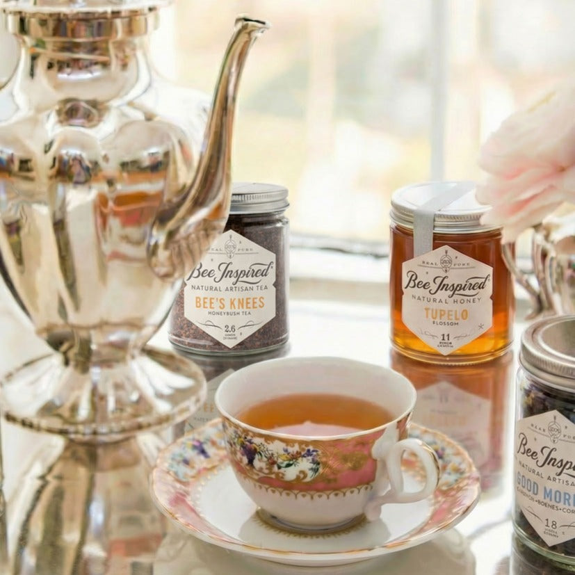 Tea cup with floral design on a saucer, surrounded by jars labeled 'Bee Inspired' on a reflective surface.