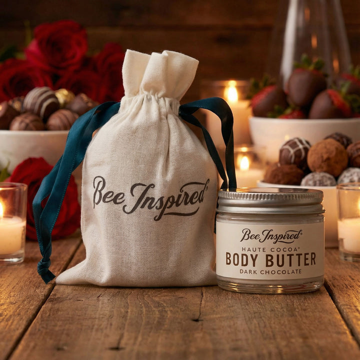Bee Inspired body butter jar and drawstring bag on a wooden surface with chocolate-covered strawberries and candles in the background.