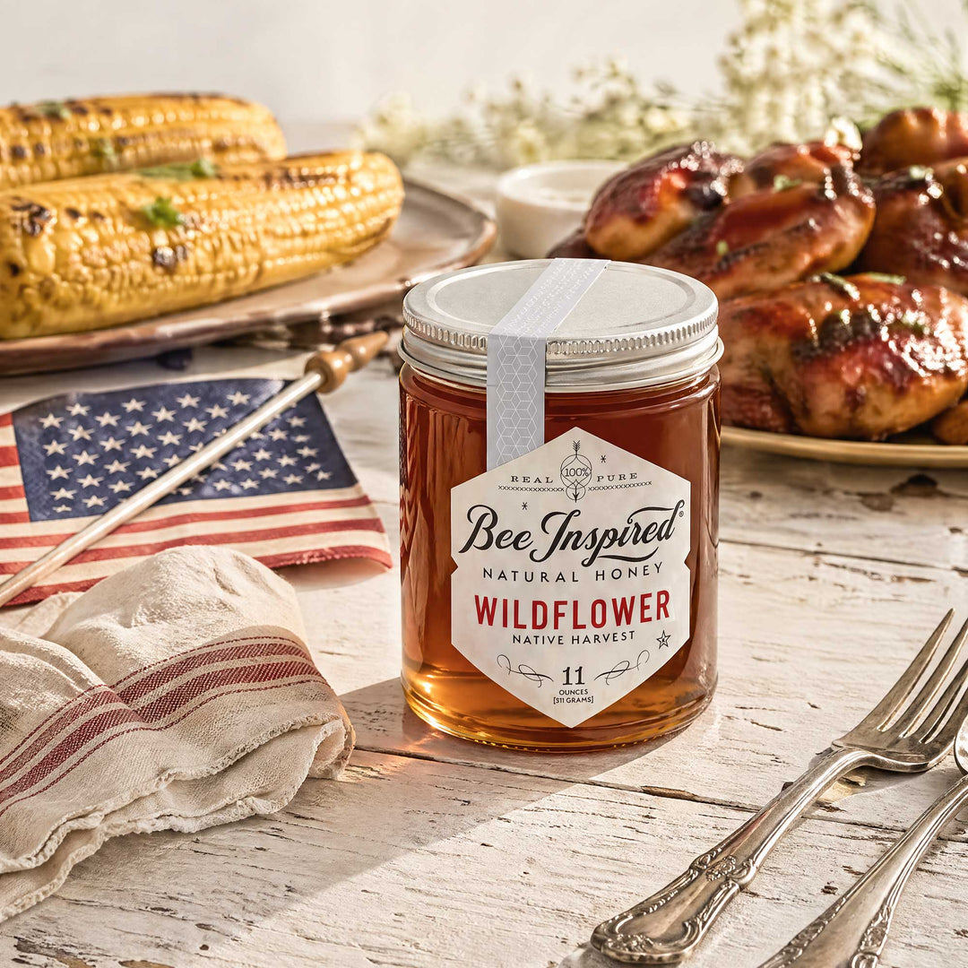 Jar of 'Bee Inspired' wildflower honey on a wooden table with grilled chicken and corn in the background.