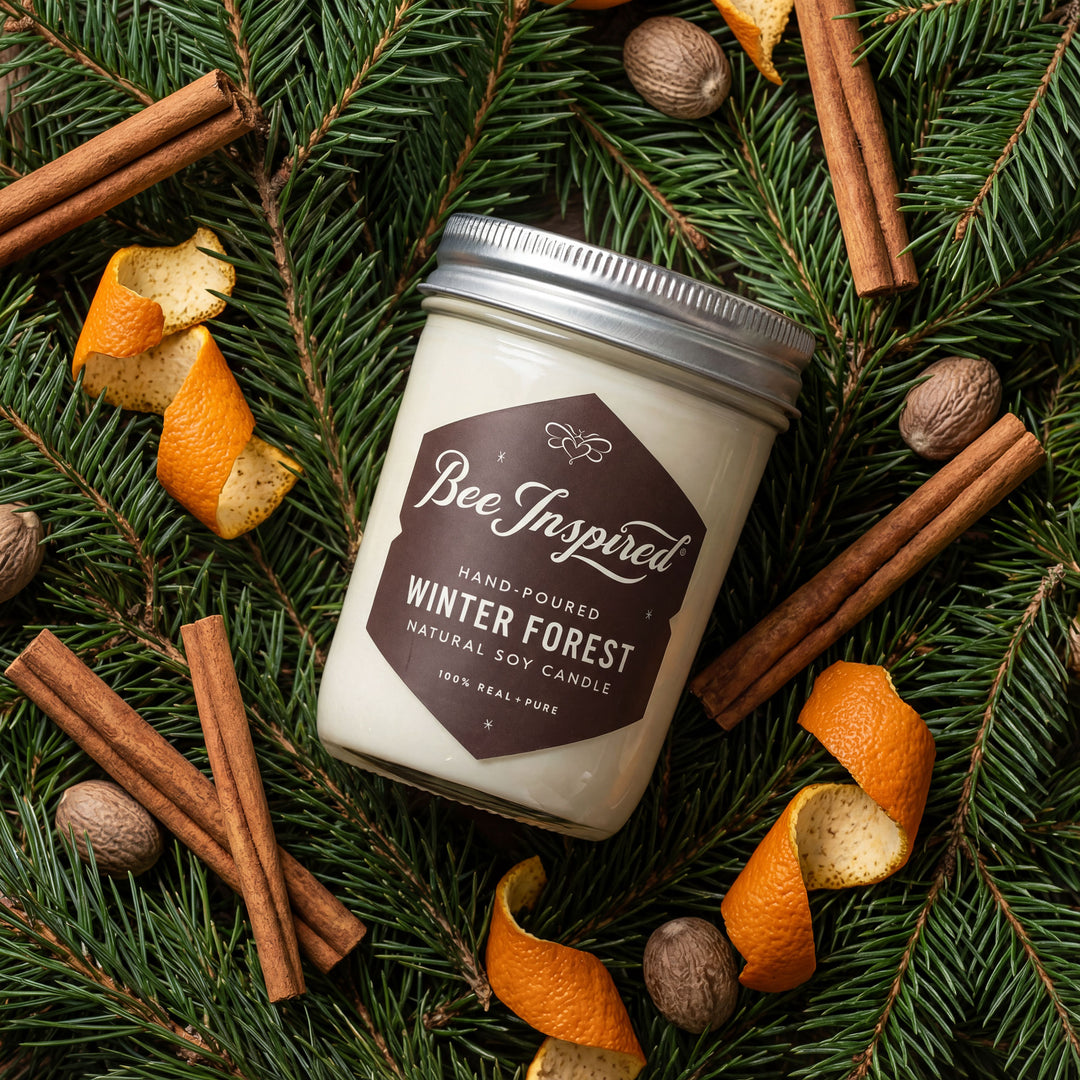 Candle labeled 'Bee Inspired' surrounded by winter elements like cinnamon sticks, oranges, and pine branches.
