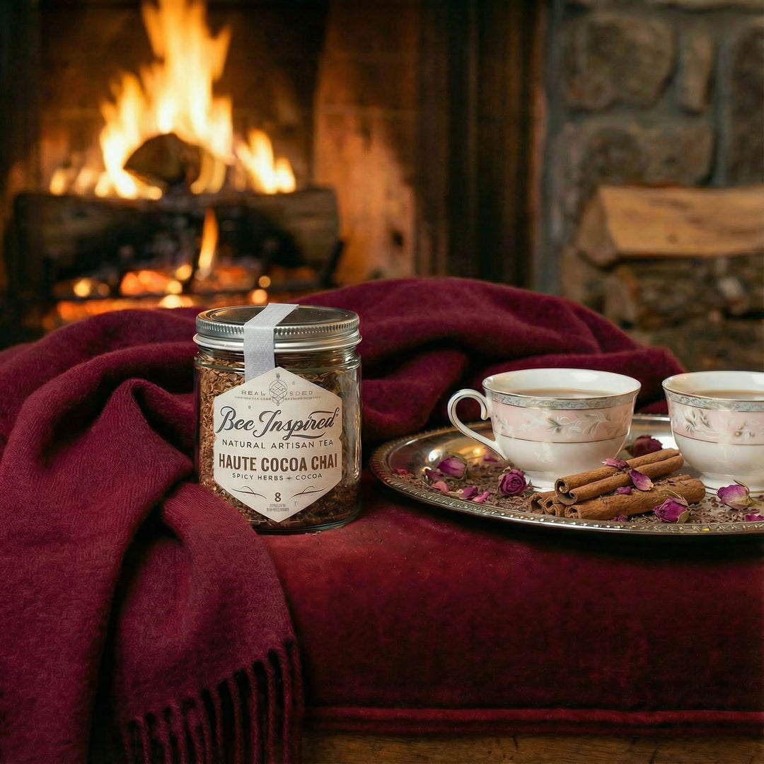 Jar of 'Bee Inspired' haute cocoa chai tea on a red blanket with teacups and cinnamon sticks in front of a fireplace.